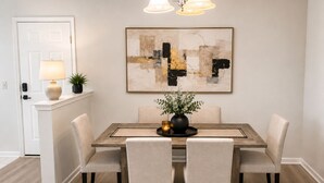 A dining area with a wooden table, four chairs, a vase with greenery, and a large abstract painting on the wall.