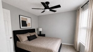 A bedroom with a bed, a ceiling fan, a nightstand with a lamp, and a window with curtains.