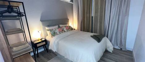 A hotel room with a bed, a nightstand, a lamp, a hanging coat rack, and a window with curtains.