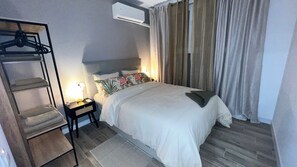 Ein Hotelzimmer mit Bett, Nachttisch, Lampe, Kleiderhaken und einem Fenster mit Vorhängen.