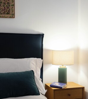 Ein ordentlich bezogenes Bett mit dunklem Kopfteil, einer Nachttischlampe und einem kleinen Nachttisch mit einem Buch.