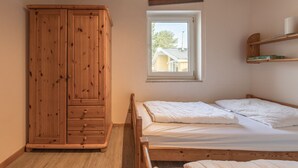 A bedroom with a wooden wardrobe, a single bed with two pillows, a window with a view of a house, and a shelf with books. - Gemutlicher Doppelbungalow mit Garten und Kaminofen in Mirow (Mirow)