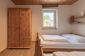 A bedroom with a wooden wardrobe, a single bed with two pillows, a window with a view of a house, and a shelf with books. - Gemutlicher Doppelbungalow mit Garten und Kaminofen in Mirow (Mirow)