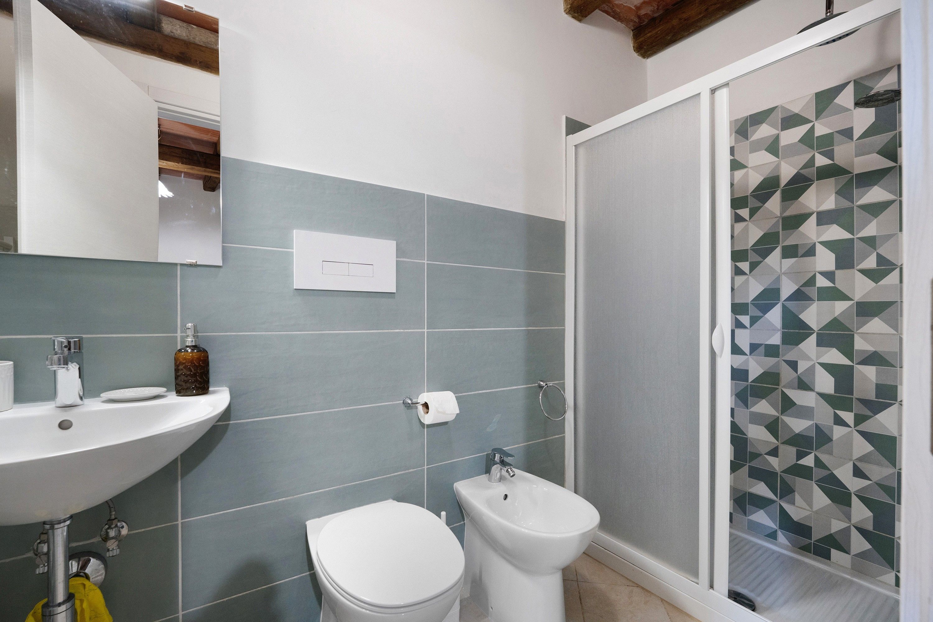 Ein Badezimmer mit Waschbecken, Toilette, Bidet und einer Dusche mit geometrischen Fliesenmustern.