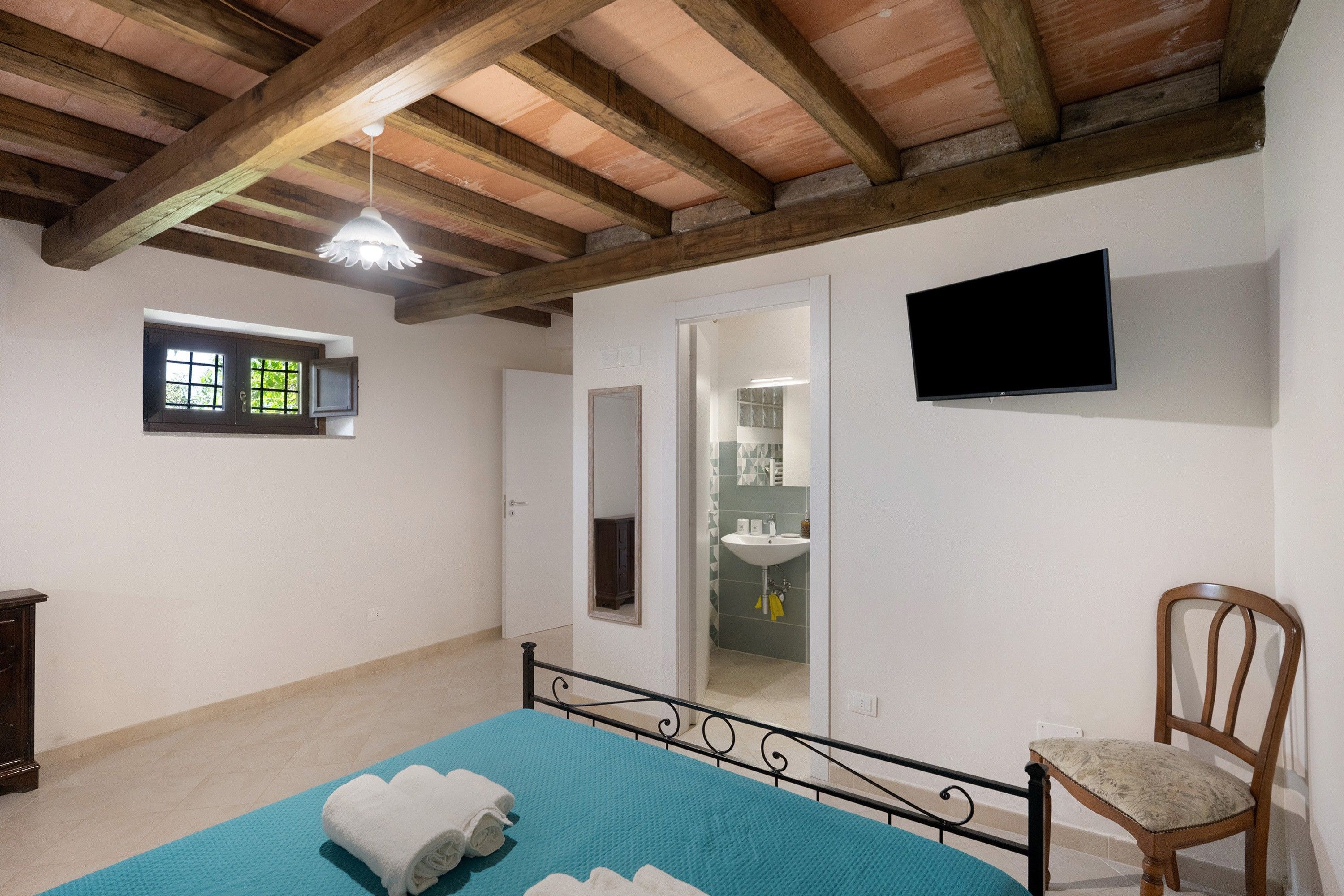 A bedroom with a bed, a chair, a television, a window, and a bathroom visible through an open door.