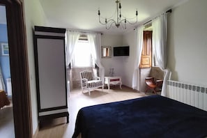 A bedroom with a bed, a chair, a small table, a TV, and a window with curtains.