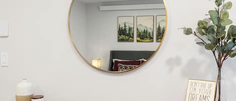 A round mirror with a gold frame, a small vase with a plant, a motivational sign, and a candle on a wooden table.