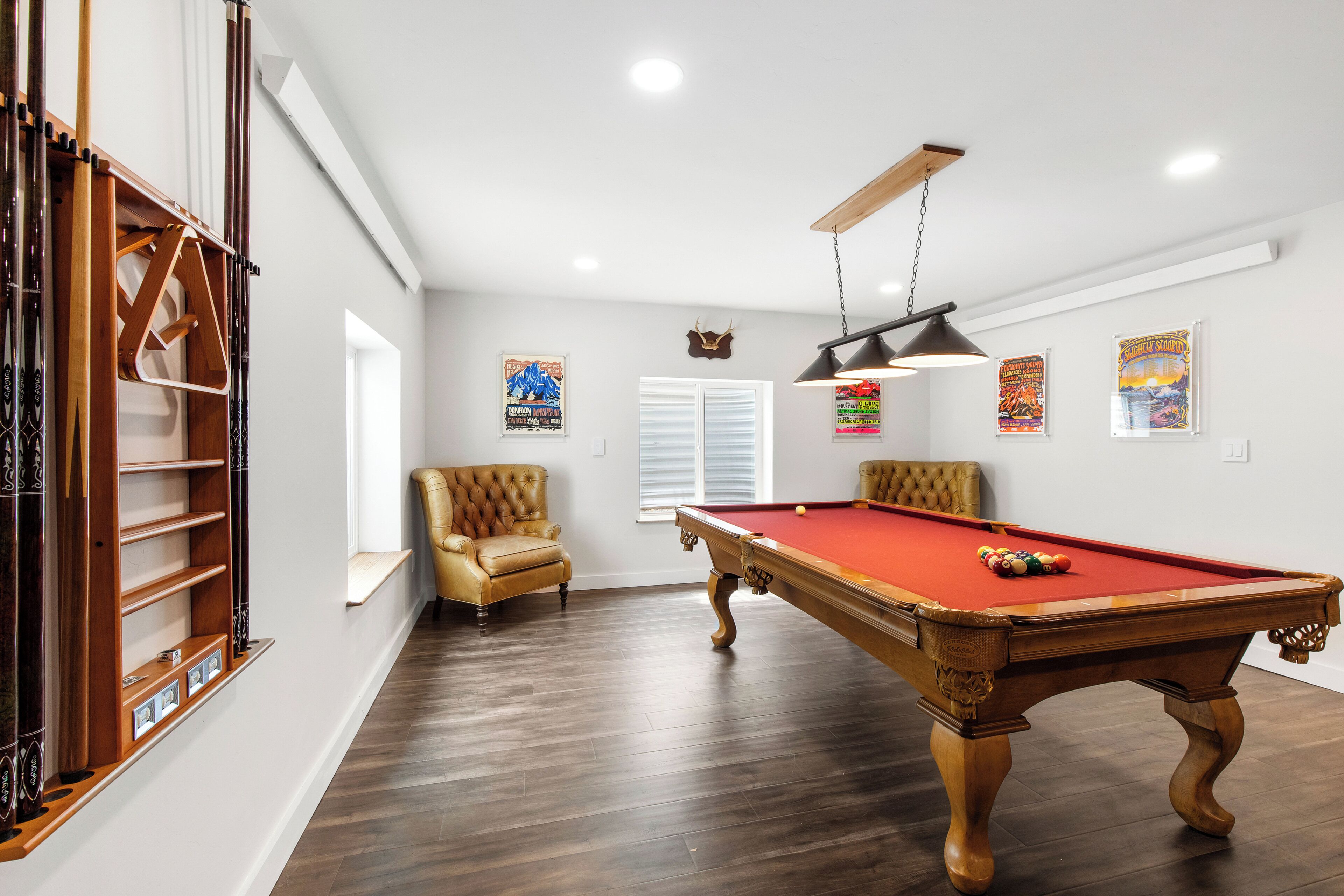 A pool table with a red felt surface, two armchairs, and a ceiling fan in a room with wooden flooring and white walls.