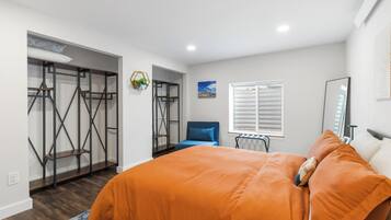 Ein Schlafzimmer mit einem Bett, einem Sessel, einem Fenster mit Jalousien und einem Kleiderschrank mit Metallregal.