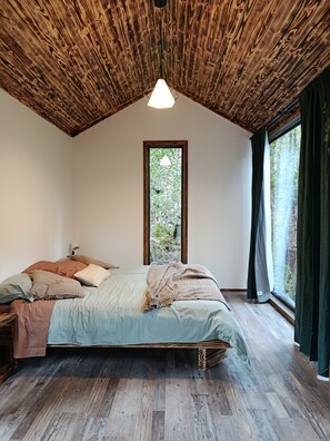 A bedroom with a sloped ceiling, a bed with multiple pillows, a window with curtains, and a view of greenery outside. - Komorebi Lodge | River, forest, clay tub, breakfast, transportation. All-inclusive (Huililco)