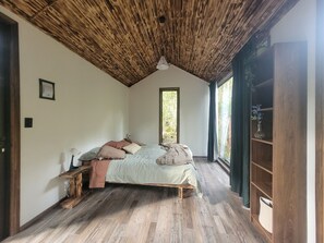 Een slaapkamer met houten plafond, een bed met kussens, een nachtkastje en een raam met uitzicht op groen.