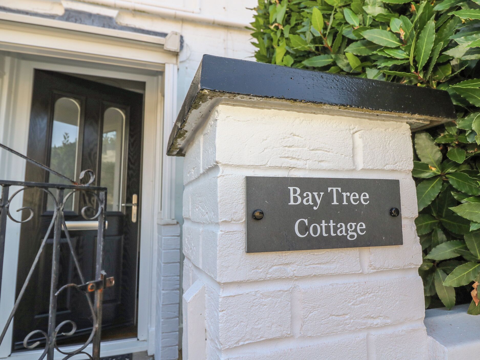 Schild für Bay Tree Cottage, ein weißes Backsteingebäude mit schwarzem Dach und einem Metalltor.