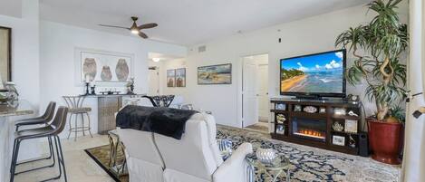 Un salon avec un canapé blanc, un téléviseur, un ventilateur de plafond et une grande plante en pot.