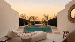A modern outdoor seating area with a pool, white cushions, a round table, and a view of palm trees and buildings in the background. - Leblanc Collection Mediterranean Villa 1 (Limmasol)