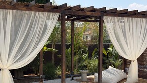 A wooden pergola with white drapes, a garden with plants, and a seating area with cushions. - Leblanc Collection Mediterranean Villa 1 (Limmasol)