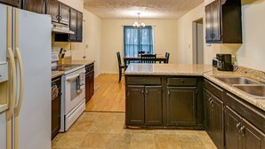 A kitchen with dark cabinets, a white refrigerator, a stove, a dining table with chairs, and a chandelier.