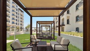 A covered outdoor seating area with tables and chairs, a grassy lawn, and a view of residential buildings. - Silkhaus Premium 1BR Stay in Yas Golf Collection (Abu Dhabi)