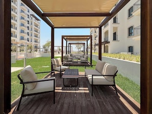 A covered outdoor seating area with tables and chairs, a grassy lawn, and a view of residential buildings. - Silkhaus Premium 1BR Stay in Yas Golf Collection (Abu Dhabi)