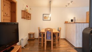 A cozy room with a wooden dining table, chairs, a television, a kitchen area with a microwave, and a wood-burning stove. - Gemutlich Eingerichteter Doppelbungalow mit Garten in Mirow (Mirow)