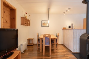 A cozy room with a wooden dining table, chairs, a television, a kitchen area with a microwave, and a wood-burning stove. - Gemutlich Eingerichteter Doppelbungalow mit Garten in Mirow (Mirow)