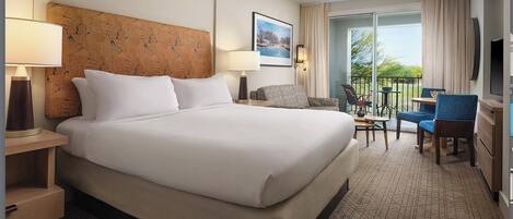 A hotel room with a large bed, a desk with a chair, a small table with a chair, and a balcony with a view.