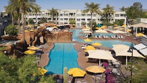 Uma área de piscina de hotel com várias piscinas, espreguiçadeiras, guarda-sóis e palmeiras.