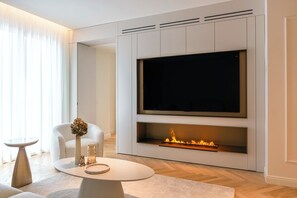 Ein modernes Wohnzimmer mit einem an einer weißen Wand befestigten Flachbildfernseher, einem Kamin darunter und einem runden Couchtisch mit Dekorationselementen.