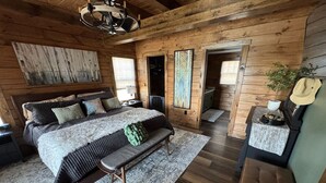 A log cabin bedroom with a large bed, a bench, a dresser, and a small table with a hat and plant. - Luxury 7BR Lakefront Log Lodge; Sleeps 18 (DuBois)