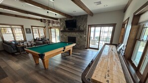 A pool table with a wooden number layout, a billiards table, and a stone fireplace in a room with wooden floors and large windows. - Luxury 7BR Lakefront Log Lodge; Sleeps 18 (DuBois)
