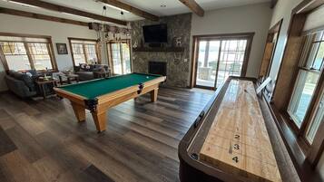 Ein Billardtisch mit Holz-Zahlenlayout, ein Spieltisch und ein Steinkamin in einem Raum mit Holzdielen und großen Fenstern.
