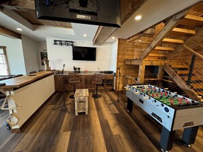 A spacious room with a wooden floor, a foosball table, a bar area, and a TV mounted on the wall. - Luxury 7BR Lakefront Log Lodge; Sleeps 18 (DuBois)