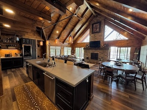 A spacious kitchen with a large island, wooden ceiling beams, and a stone fireplace. - Luxury 7BR Lakefront Log Lodge; Sleeps 18 (DuBois)