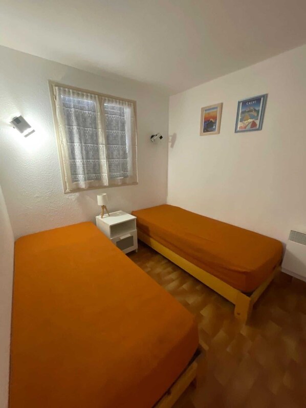 A room with two beds, a small white bedside table, a window with patterned curtains, and two wall-mounted pictures.