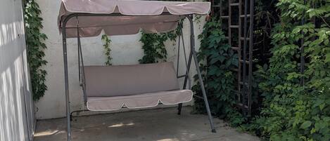 Uma cadeira balanço de metal com toldo, posicionada em frente a uma parede coberta por plantas trepadeiras.
