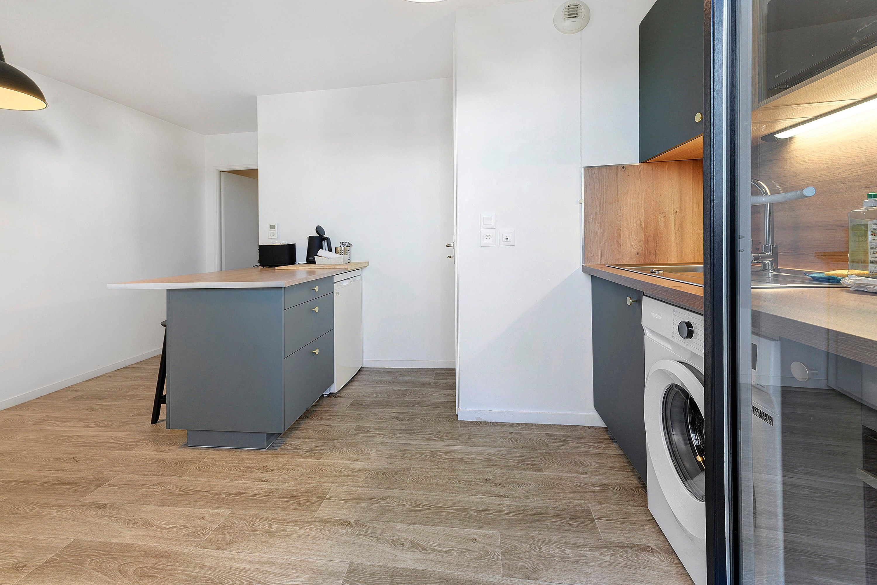 Une cuisine compacte dotée d’un lave-linge encastré, d’un petit îlot central avec un évier et d’un plan de travail en bois.