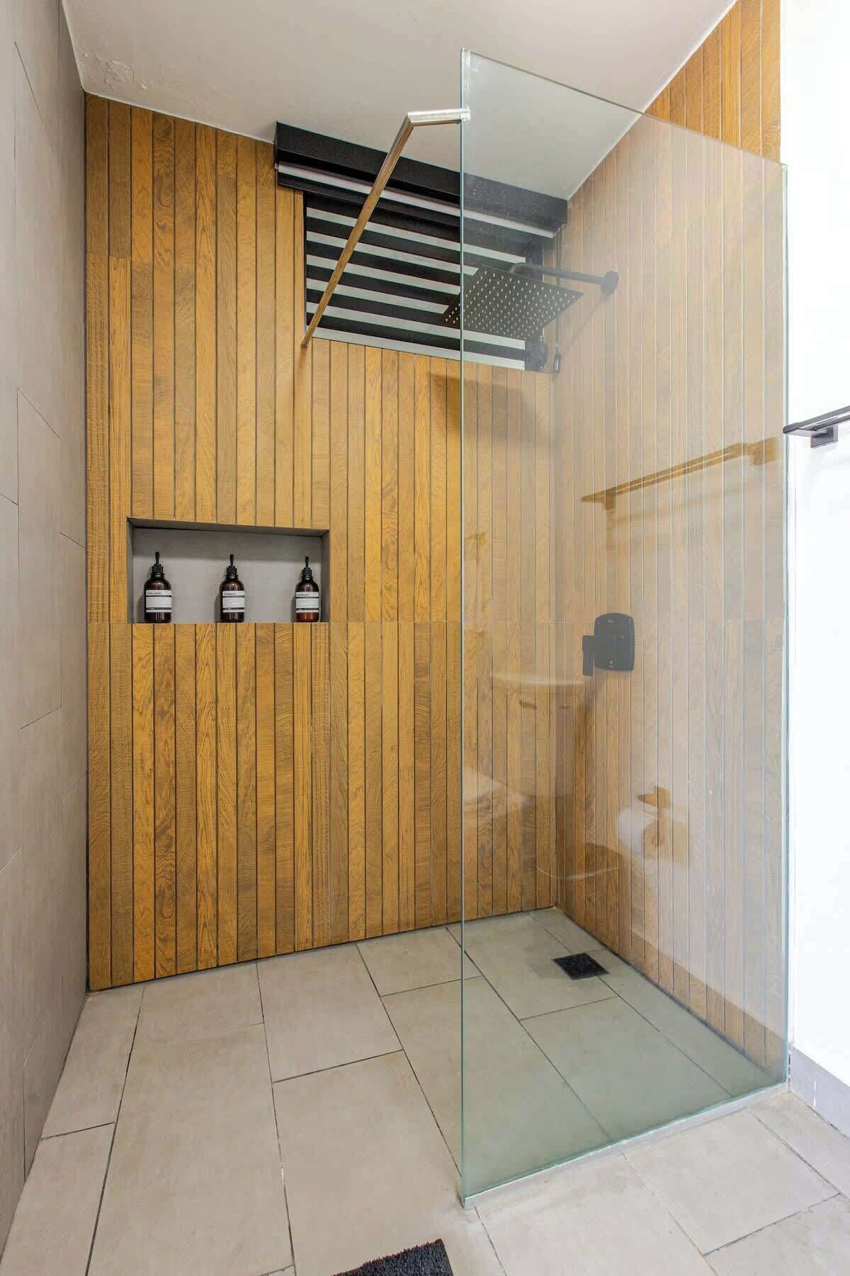 Un baño moderno con paneles de madera, mampara de vidrio y estante con botellas.