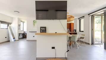 Un interior moderno con un televisor de pantalla plana montado en una pared, un estante de madera con un cartel, una zona de comedor con sillas y un amplio ventanal.