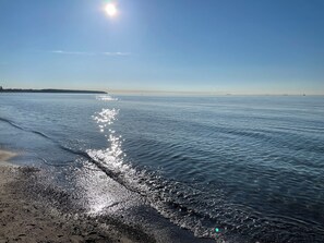 Une plage avec un ciel dégagé, une eau calme qui reflète la lumière du soleil et des reliefs lointains.