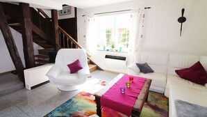 Sala de estar con un sofá blanco, un sillón blanco, una mesa de centro de madera y un tapete colorido. Se ve una ventana con cortinas y vistas al exterior con vegetación. En el fondo, se distingue una escalera.