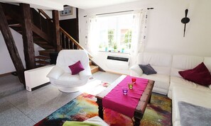 A living room with a white sofa, a white armchair, a wooden coffee table, and a colorful rug. There is a window with curtains and a view of greenery outside. A staircase is visible in the background. - Gestrandet Vacation Home | Quiet Location | 20 Min. to the Baltic Sea | Sleeps up to 4 (Satow)