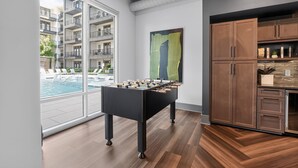 Un interior moderno con mesa de fútbol, armarios de madera y vista a una piscina y edificios de apartamentos.