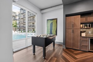 Ein modernes Interieur mit Kicker, Holzschränken und Blick auf einen Pool sowie Apartmentgebäude.