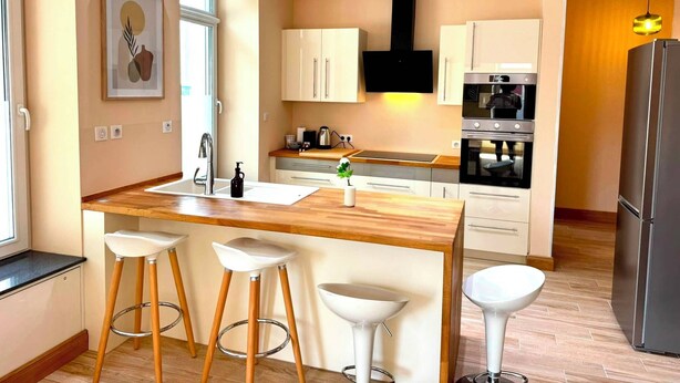 Une cuisine moderne dotée d’un plan de travail en bois, de tabourets de bar blancs, d’un four encastré et d’un réfrigérateur en acier inoxydable.