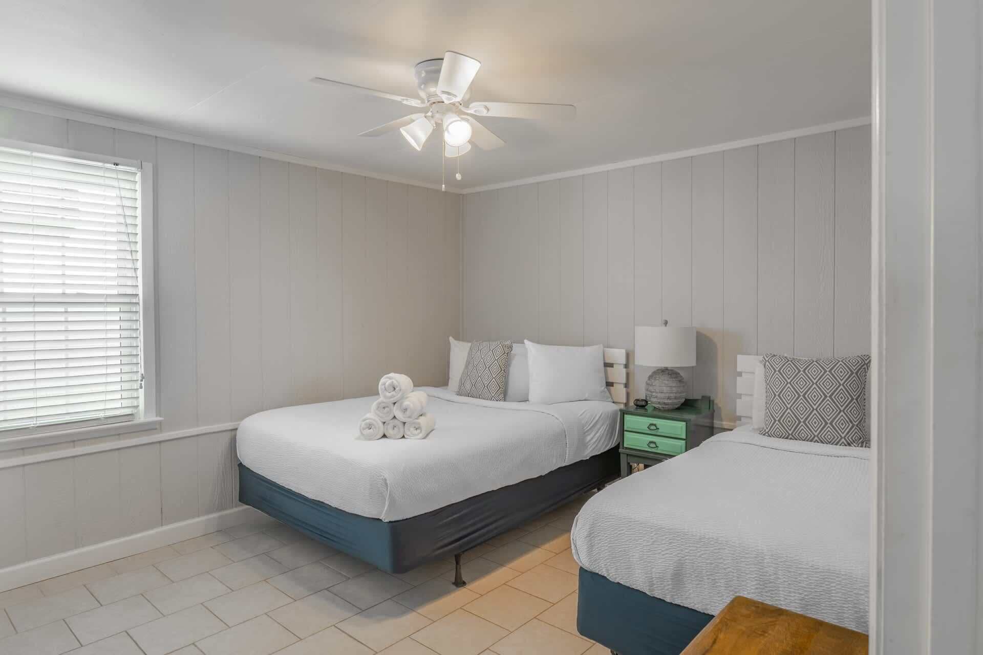 A bedroom with two beds, a ceiling fan, a nightstand with a lamp, and a window with blinds.