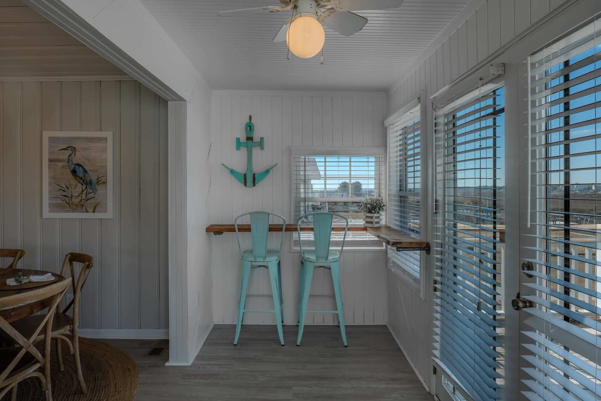 A room with a wooden dining table, two chairs, a ceiling fan, a window with blinds, and a wall-mounted anchor decoration.