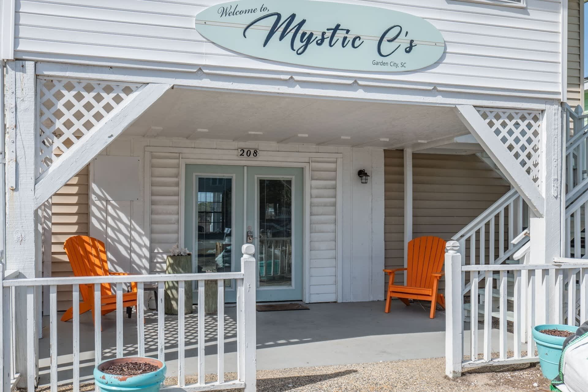 Un porche avec deux fauteuils Adirondack orange, une boîte aux lettres et une pancarte indiquant : « Mystic C's Garden City, Caroline du Sud ».
