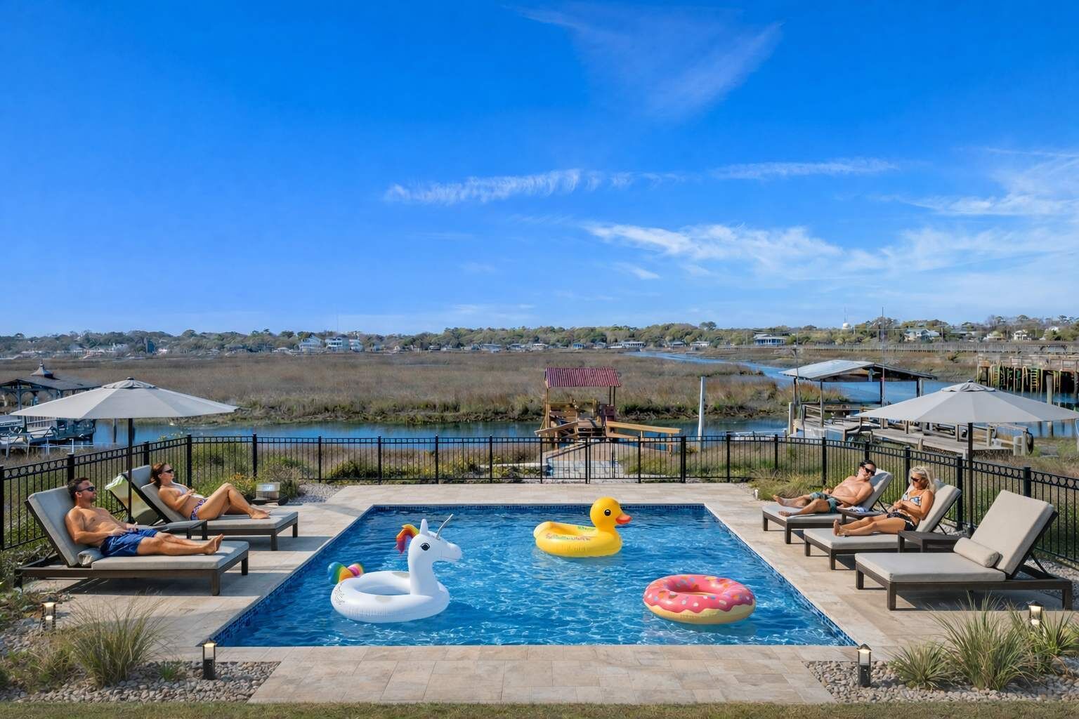 Ein Poolbereich mit Liegestühlen und aufblasbaren Spielzeugen, umzäunt und mit Blick auf ein Gewässer.