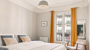Ett hotellrum med en säng, en stol, ett fönster med gardiner, en tavla på väggen och en taklampa.