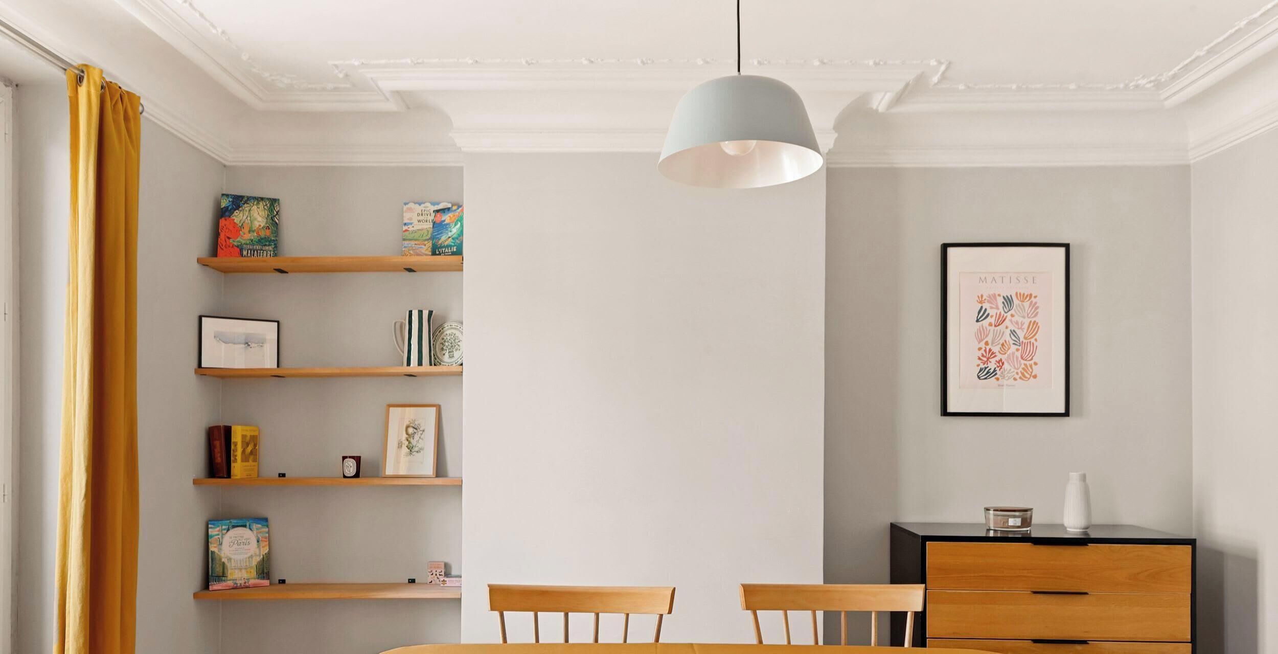 A dining area with a table, chairs, a pendant light, and a bookshelf with books and decorative items.