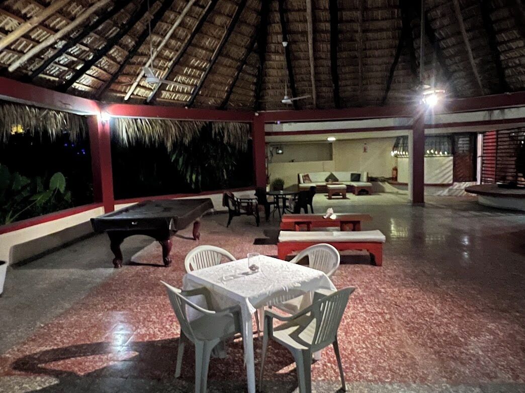 Un espacio al aire libre con mesas y sillas, una mesa de billar y una estructura con techo de paja.